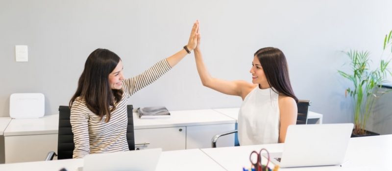 Smiling female professionals giving high-five after completing tasks at desk in workplace